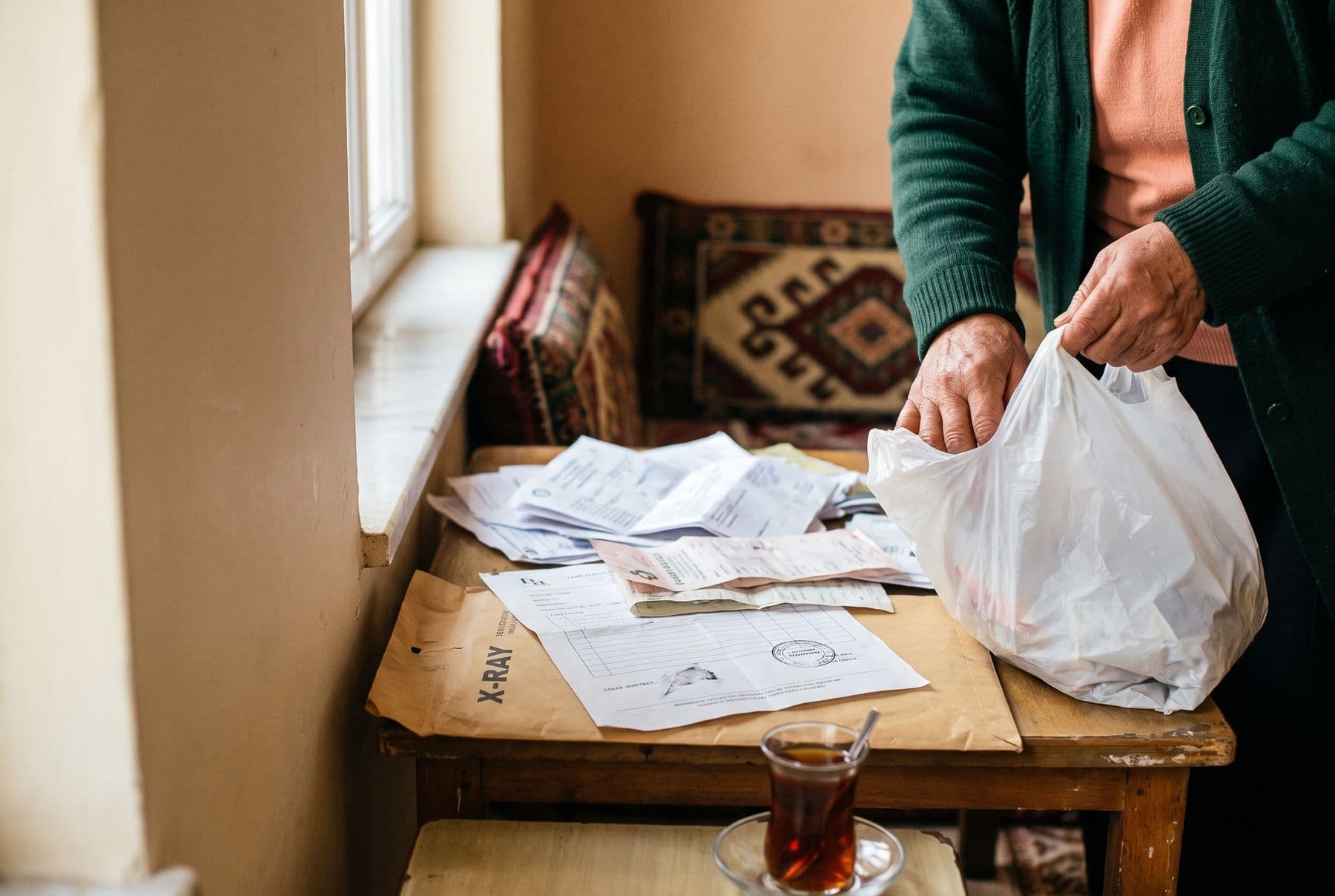 An older person tipping a plastic bag of prescriptions and lab papers onto a wooden table, next to an X-ray envelope and a glass of tea.